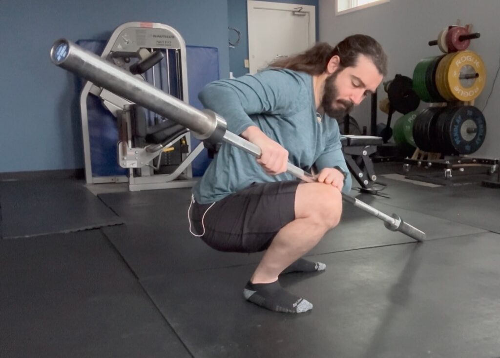 Dr. John using the weight of a barbell to press against his inner thigh in a squat position as a pre squat warm up